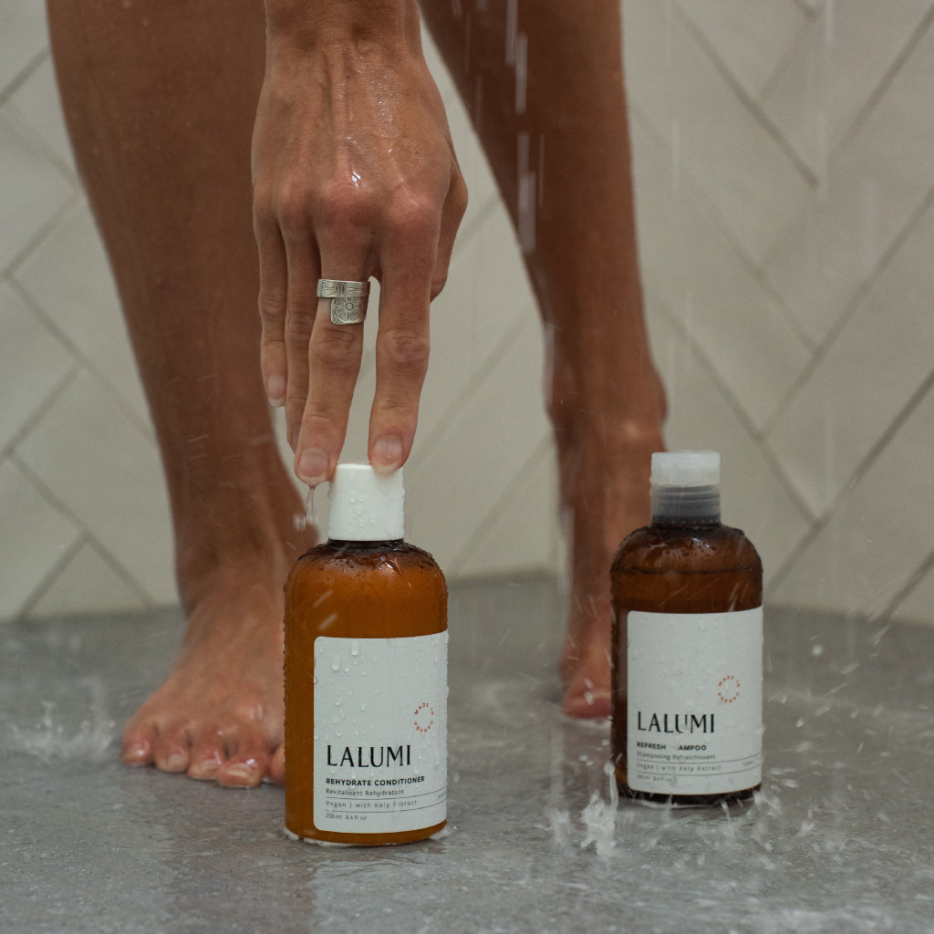 Image of woman reaching down to pick up lalumi Rehydrate Conditioner besides Lalumi Refresh Shampoo in Shower
